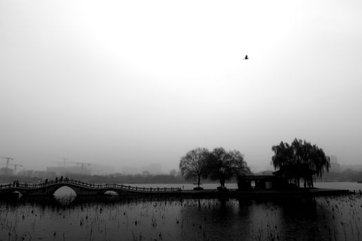 Serene black and white landscape with a bridge over a lake and trees in the mist.