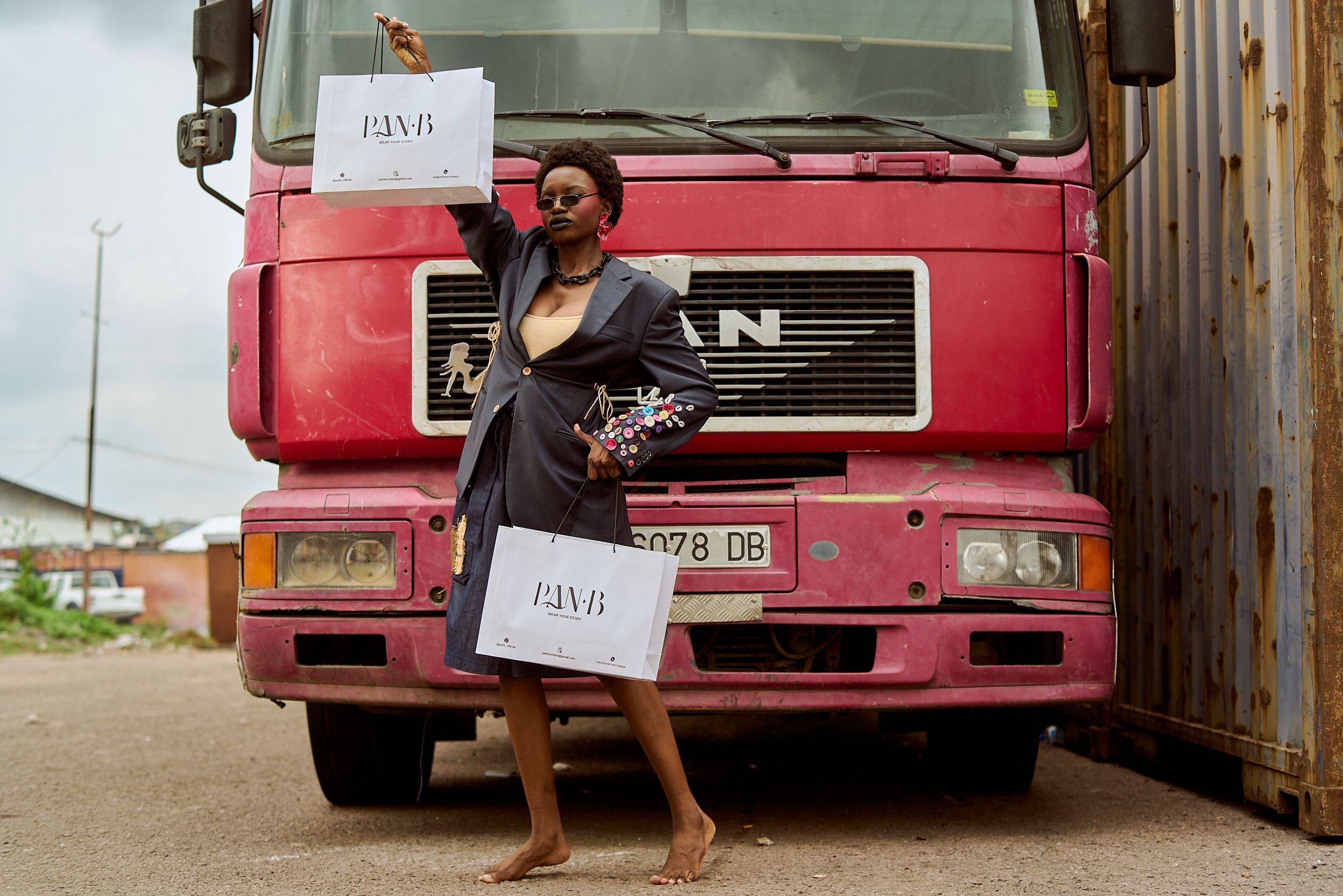 Stylish woman posing barefoot with shopping bags in front of a pink truck outdoors.