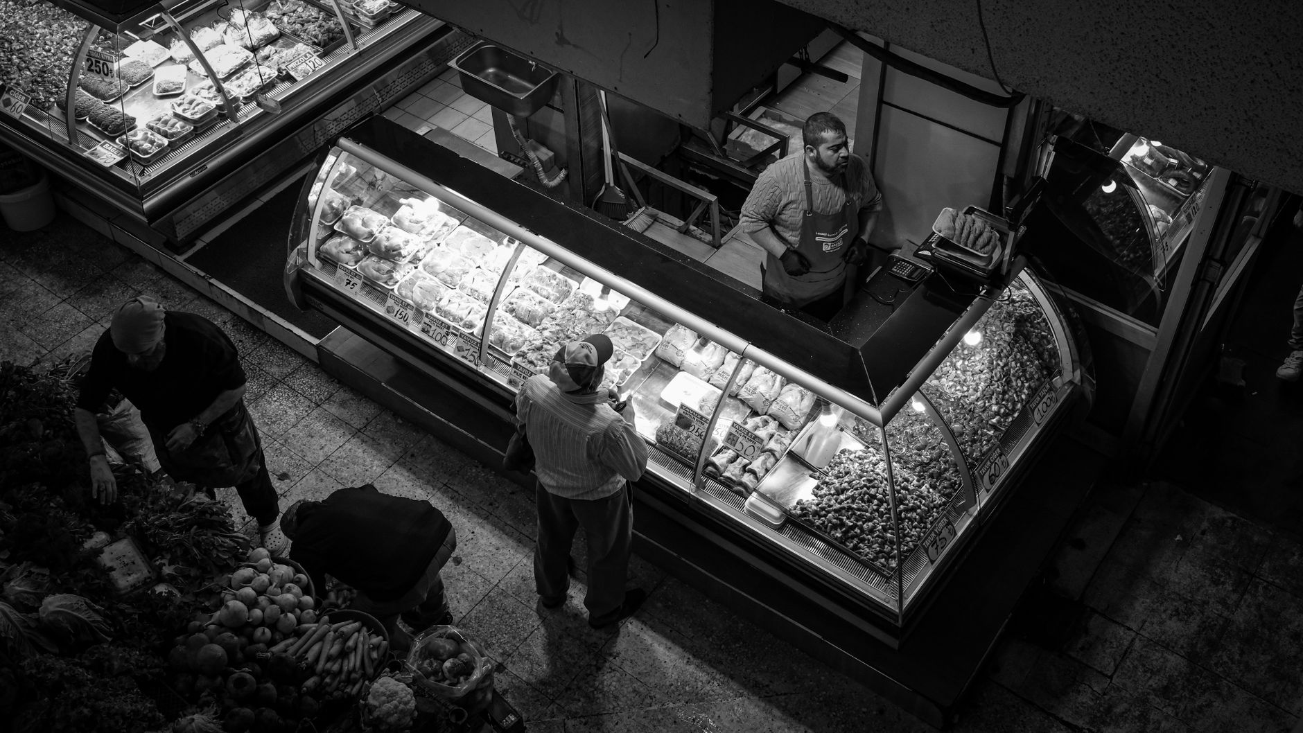 A vibrant market scene with a vendor engaging customers at a deli counter, captured in black and white.