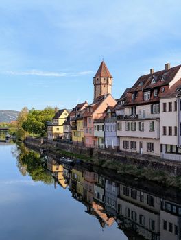 Picturesque European village with colorful houses reflected in calm river water under blue sky.