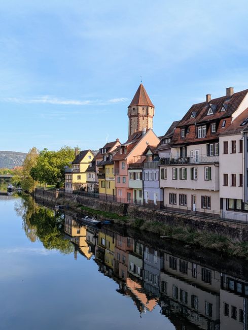 Picturesque European village with colorful houses reflected in calm river water under blue sky.