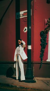 Fashionable woman in white suit poses by a street lamp against a red wall, exuding elegance.