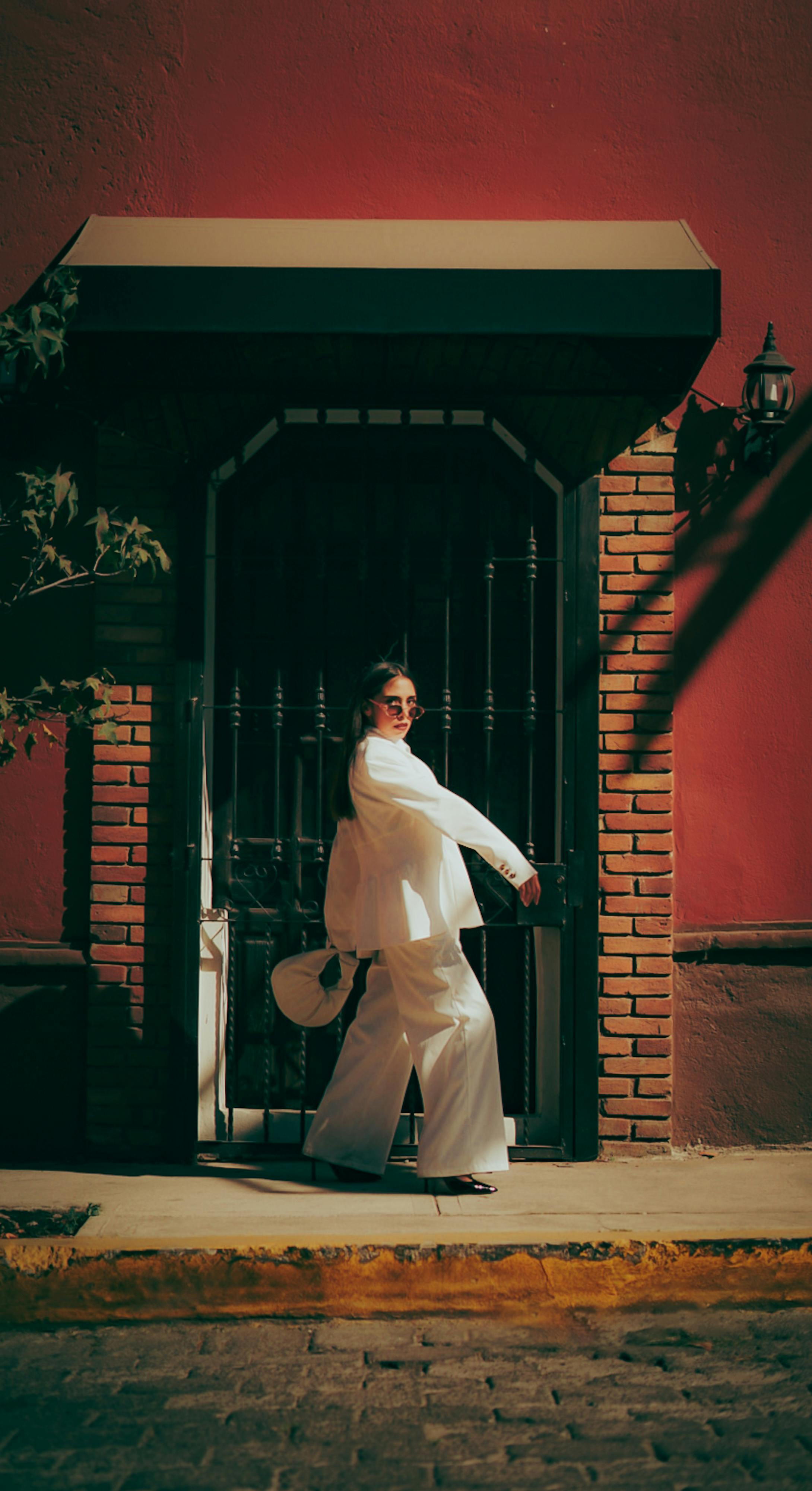 бесплатная В солнечный день мимо ярко-красной стены проходит модная женщина в белом костюме. Стоковое фото