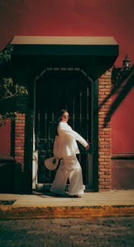 Fashionable woman in a white suit walking past a vibrant red wall on a sunny day.