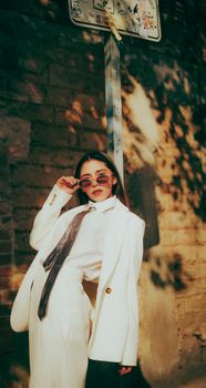 Fashionable woman in a white suit posing with sunglasses against a brick wall in warm sunlight.
