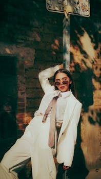 Stylish woman in white suit and sunglasses posing confidently in an urban environment with dramatic shadows.