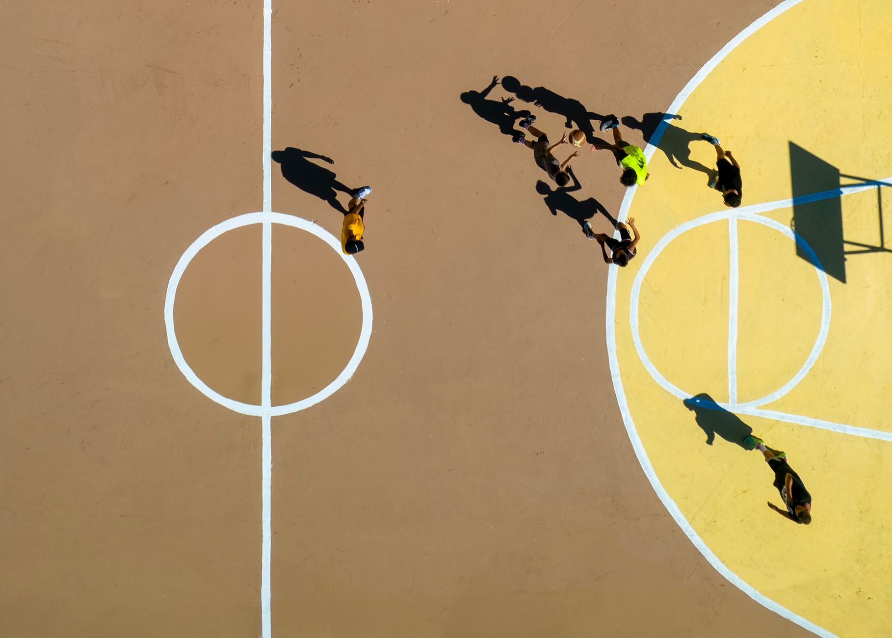 Group of adults playing basketball on an outdoor court viewed from above, showcasing dynamic motion and shadows.