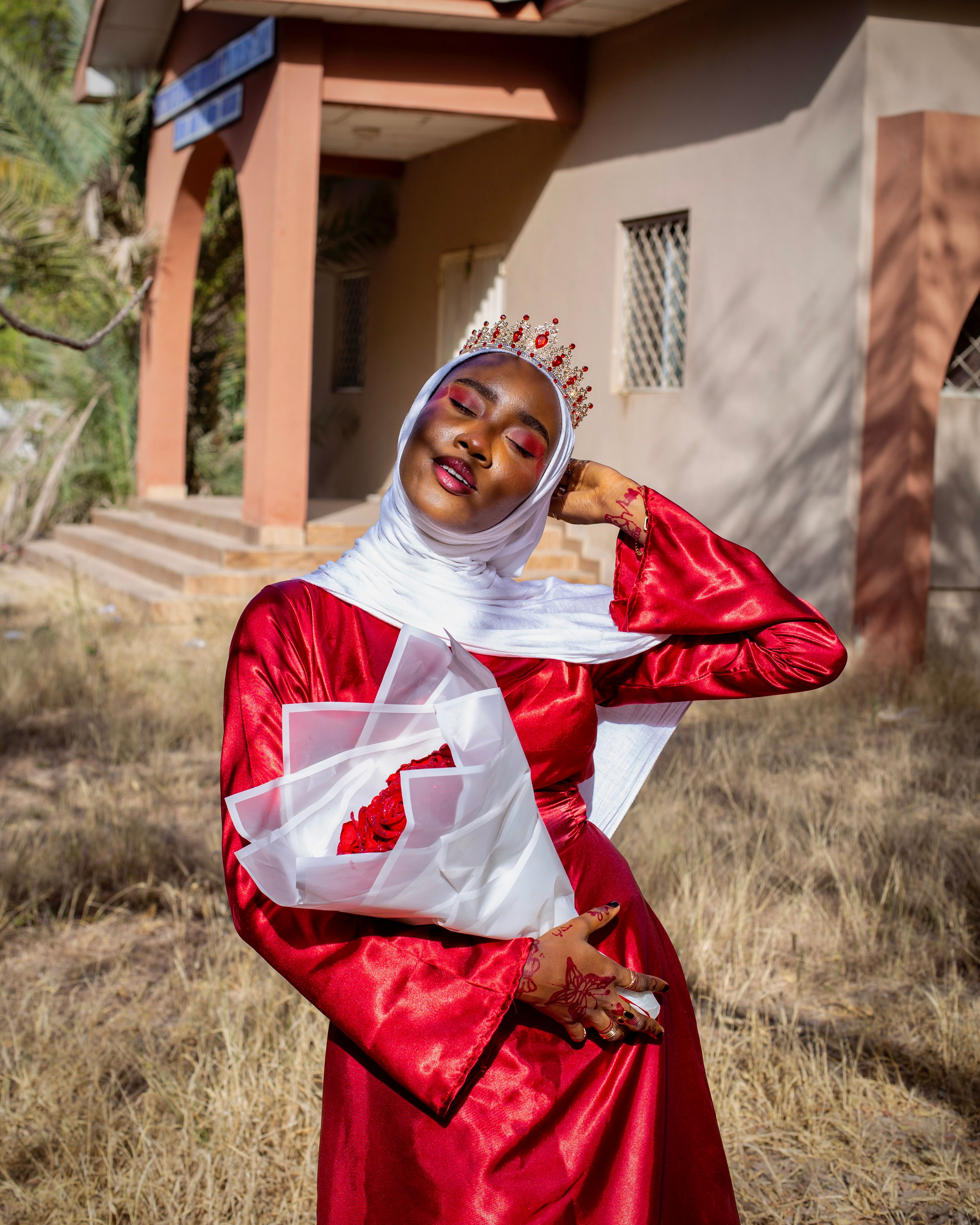 Stylish young woman in red dress and hijab with floral bouquet, enjoying sunny day outside.