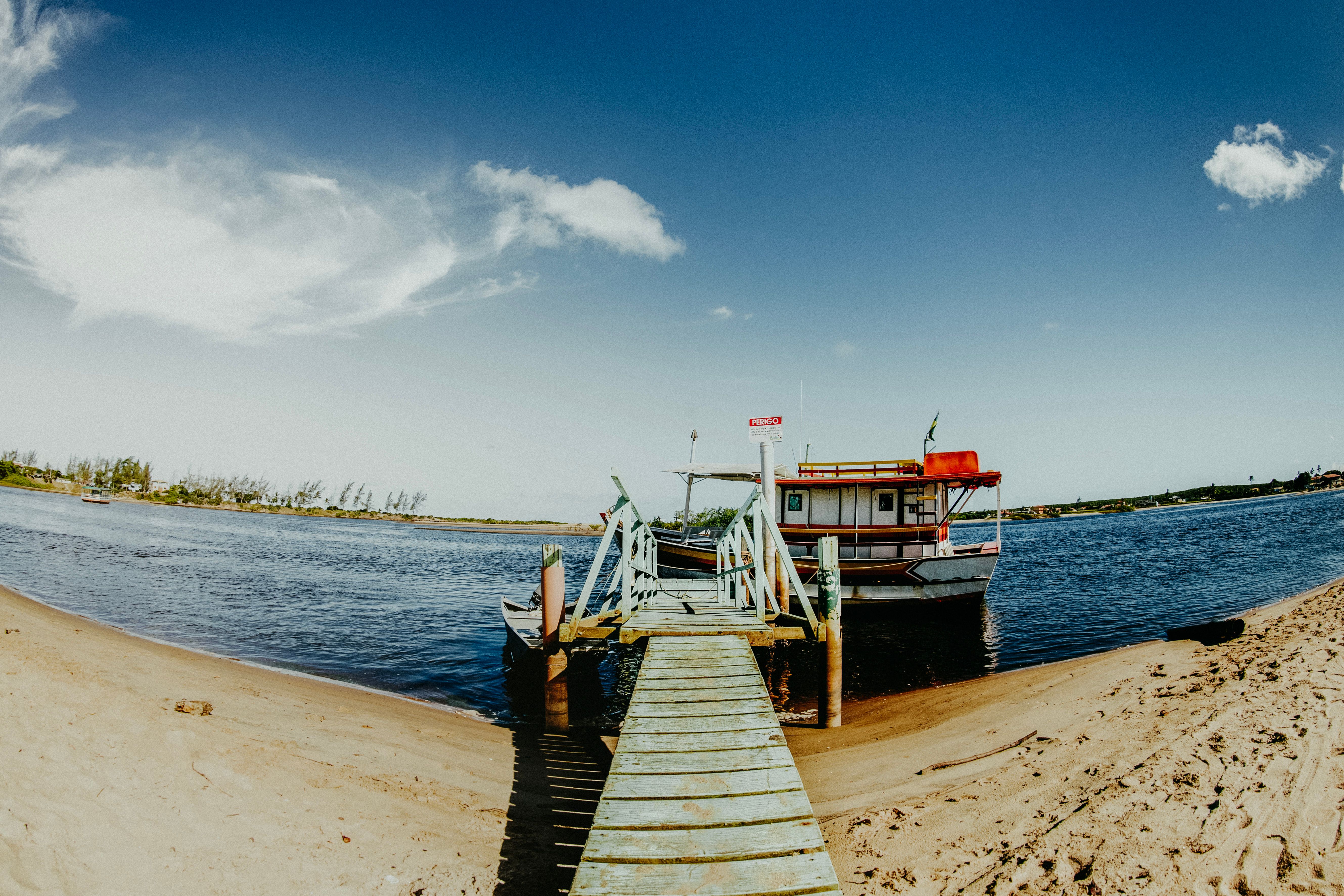 A rustic wooden pier leads to a docked boat on a serene sandy riverbank under a vivid blue sky.