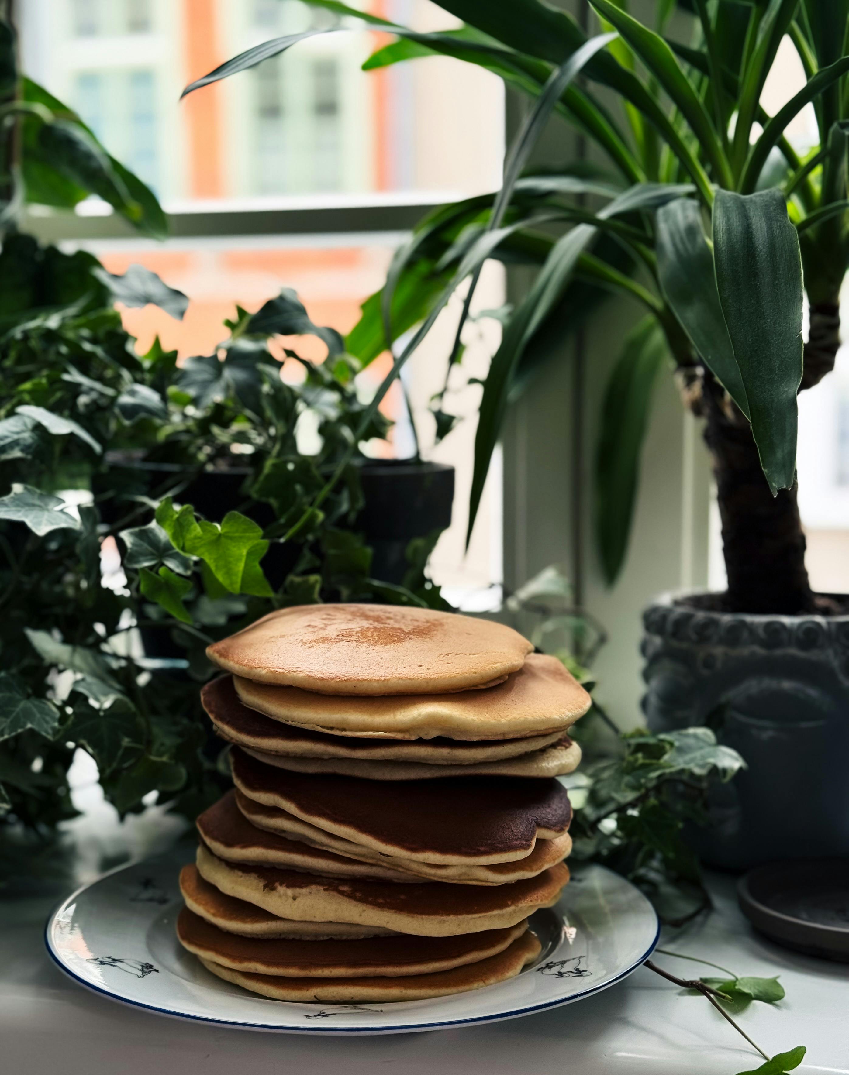 бесплатная Высокая стопка золотистых блинчиков рядом с пышными комнатными растениями у окна. Стоковое фото