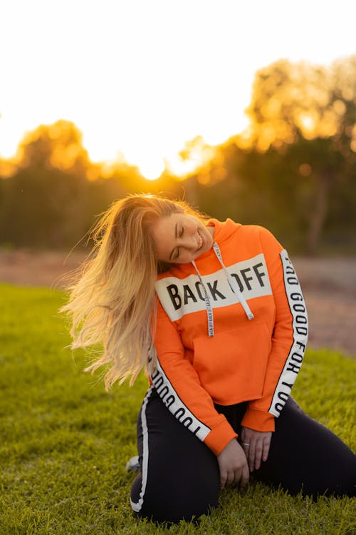 Free Woman In Orange And White Hoodie And Black Pants Kneeling On Green Grass Field Stock Photo