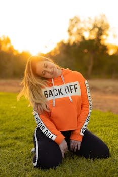 一位身穿橙色连帽衫、帽衫上写着“退后”字样的金发女子，在阳光明媚的户外享受着黄金时段的时光