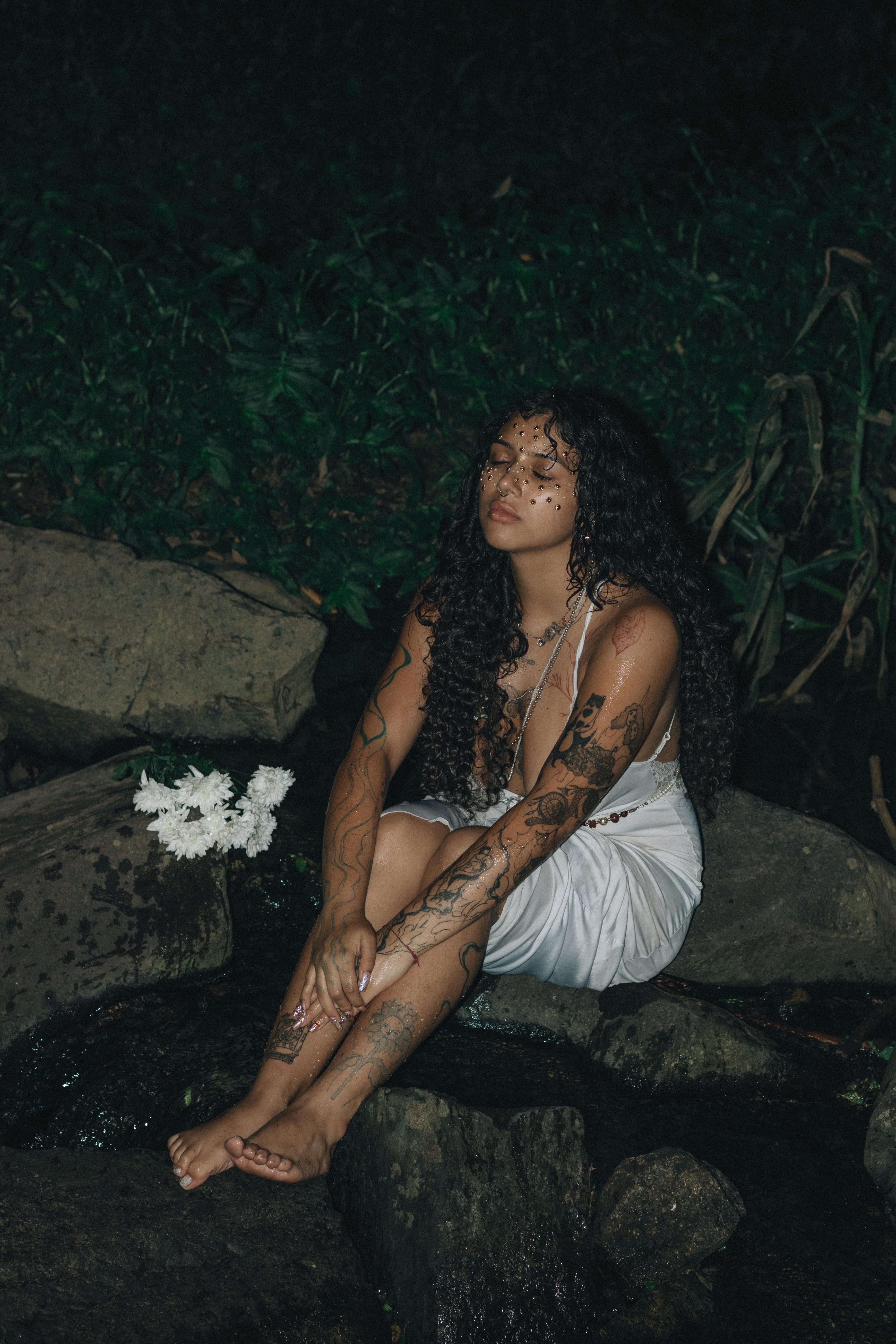 бесплатная Женщина в белом платье с татуировками сидит на камнях у ночного ручья. Стоковое фото