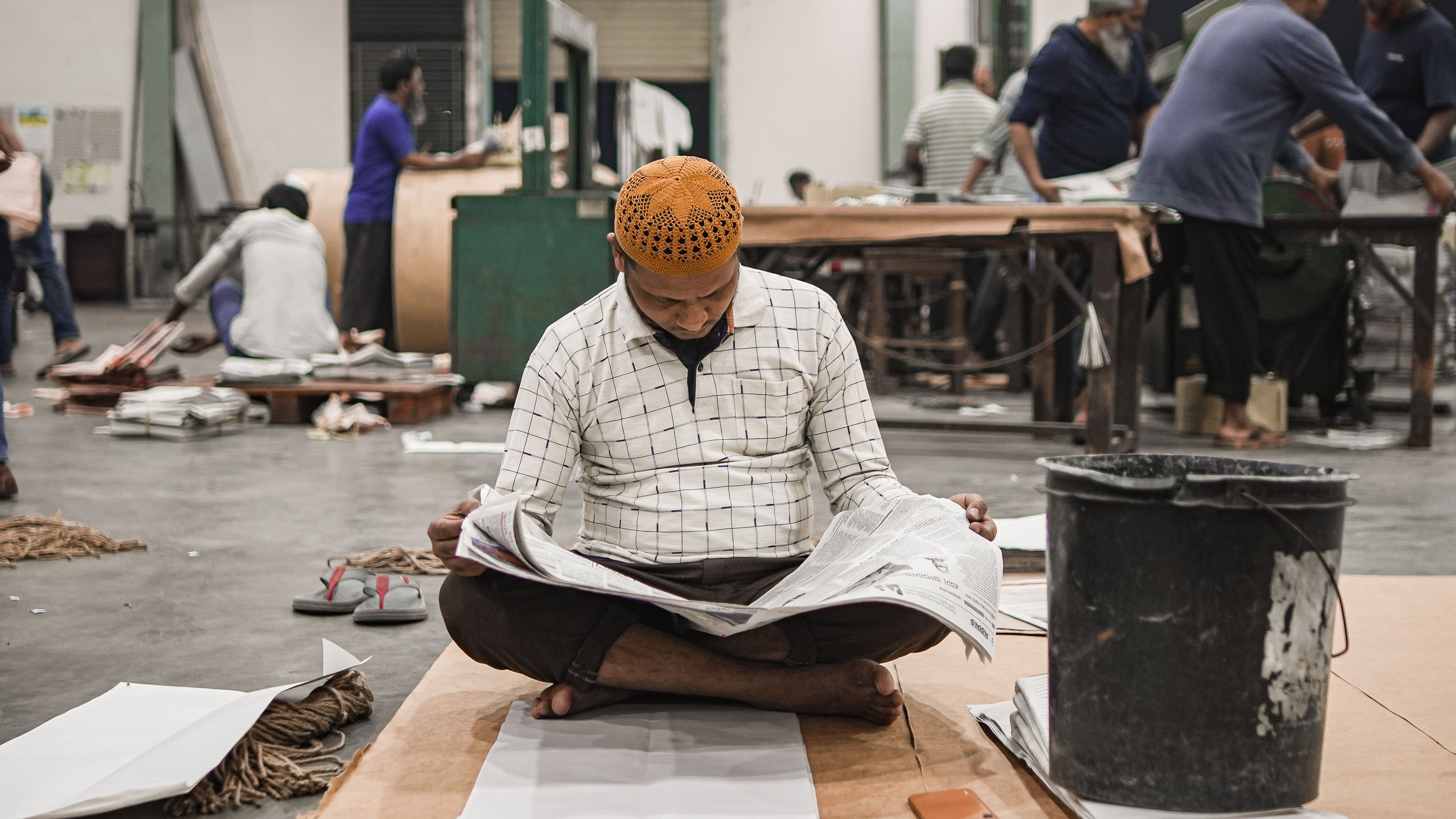 Adult man reading a newspaper in a bustling workshop setting, surrounded by busy workers and industrial equipment.