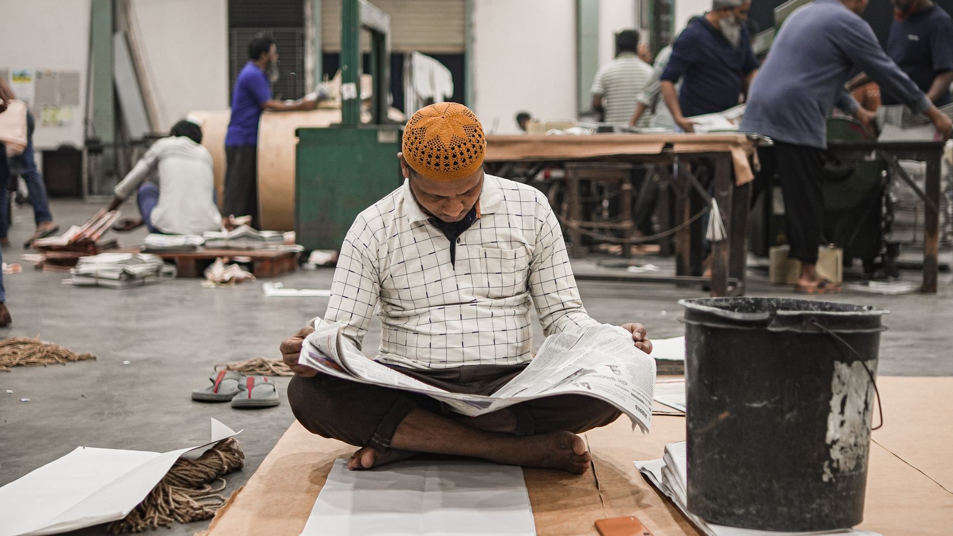 Adult man reading a newspaper in a bustling workshop setting, surrounded by busy workers and industrial equipment.