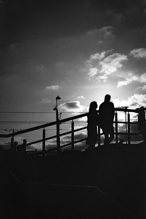 Free A romantic silhouette of a couple on a bridge with dramatic cloudy skies in the background. Stock Photo
