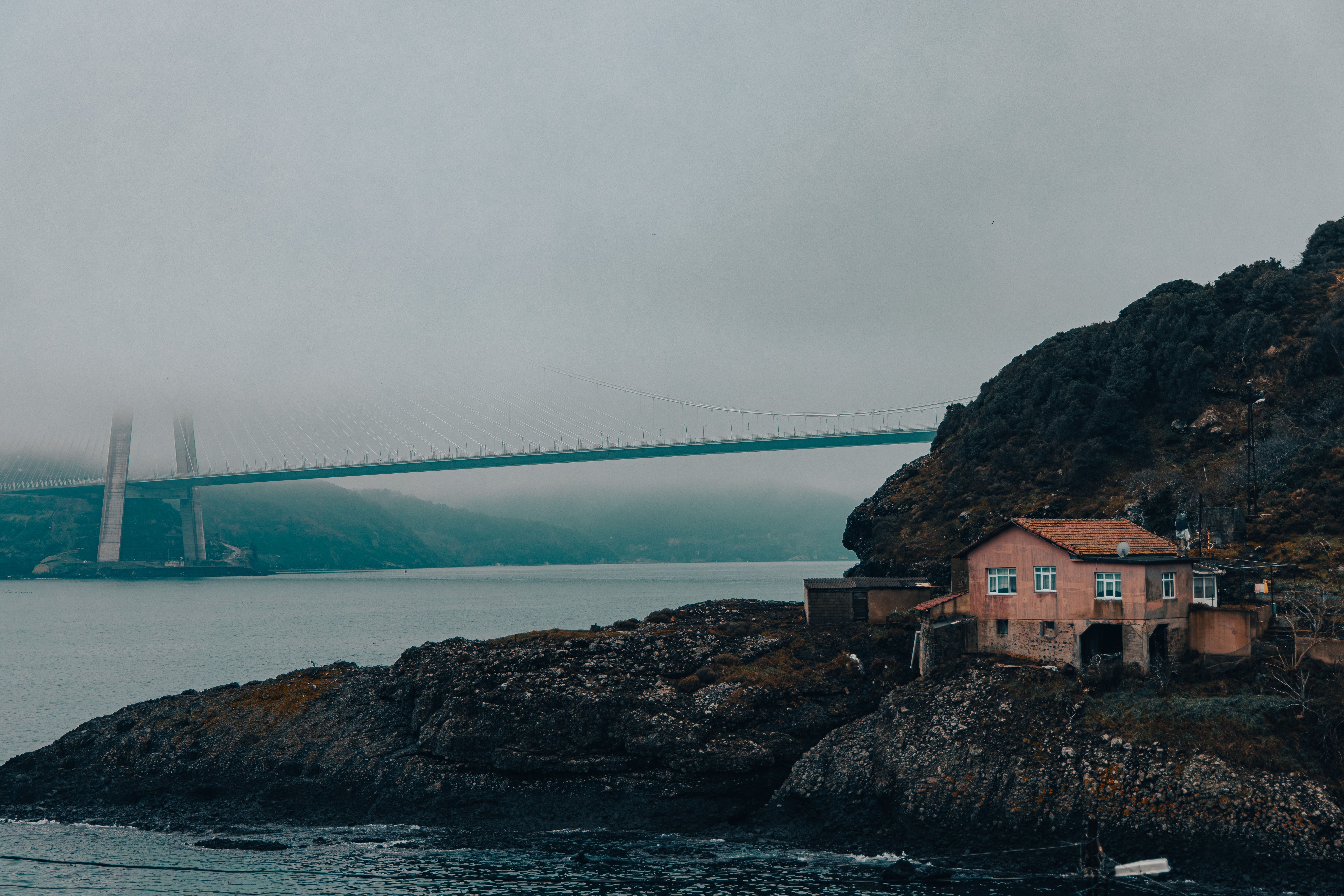 Un pont brumeux enjambe des eaux calmes, une maison côtière se dressant au premier plan, créant une scène paisible.