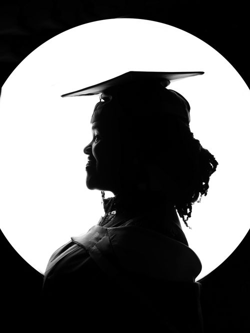 Free Artistic silhouette of a graduate wearing cap and gown with a dramatic spotlight. Stock Photo