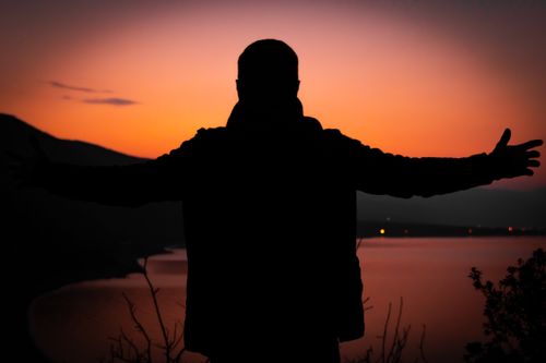 Free A person in silhouette with arms outstretched at sunset over a calm lake, creating a dramatic scene. Stock Photo