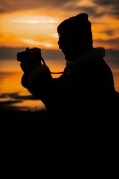 Silhouette of a person taking photos at sunset near calm waters, exuding a peaceful, artistic vibe.