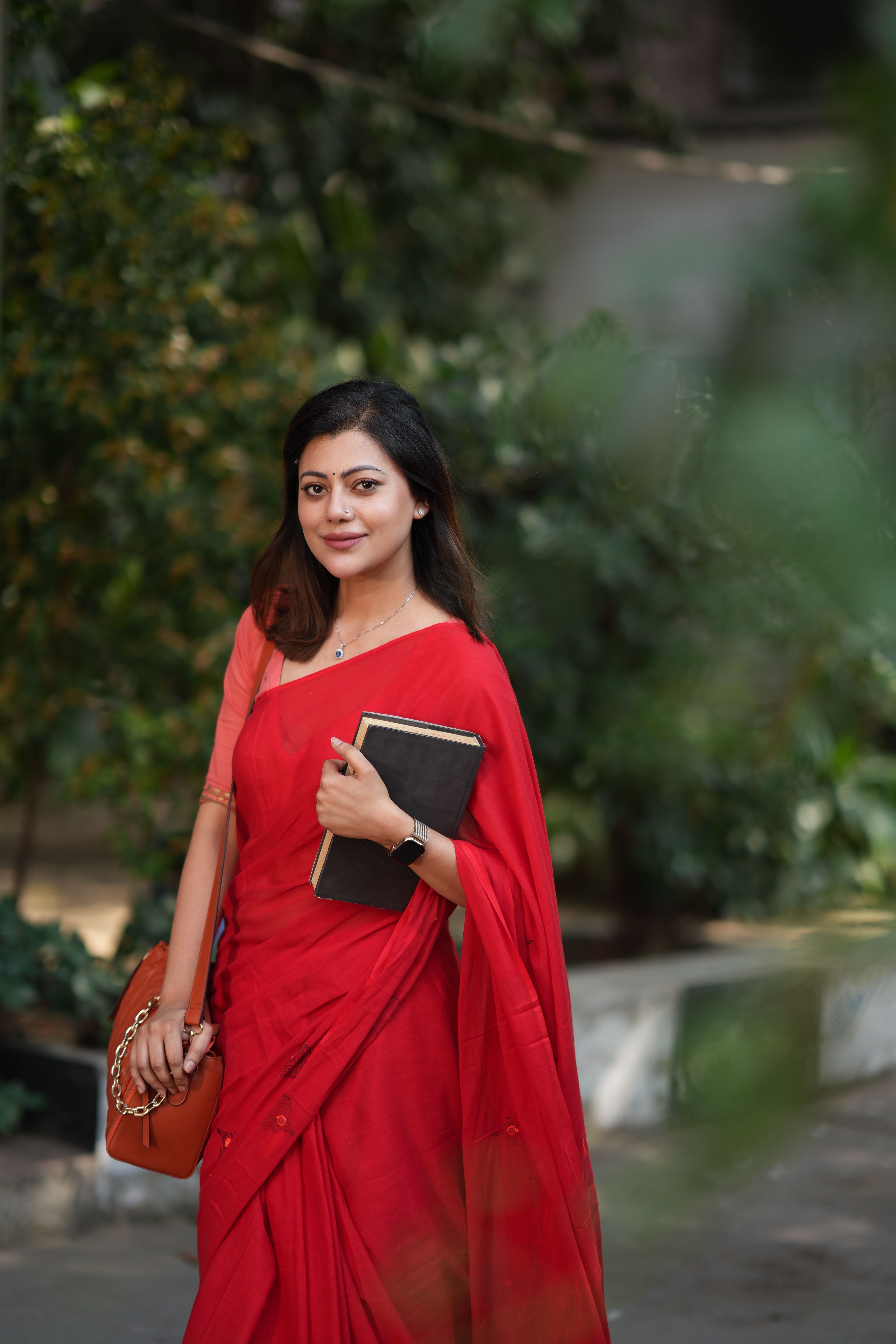 Smiling woman in red sari with a book and handbag, walking confidently outdoors.