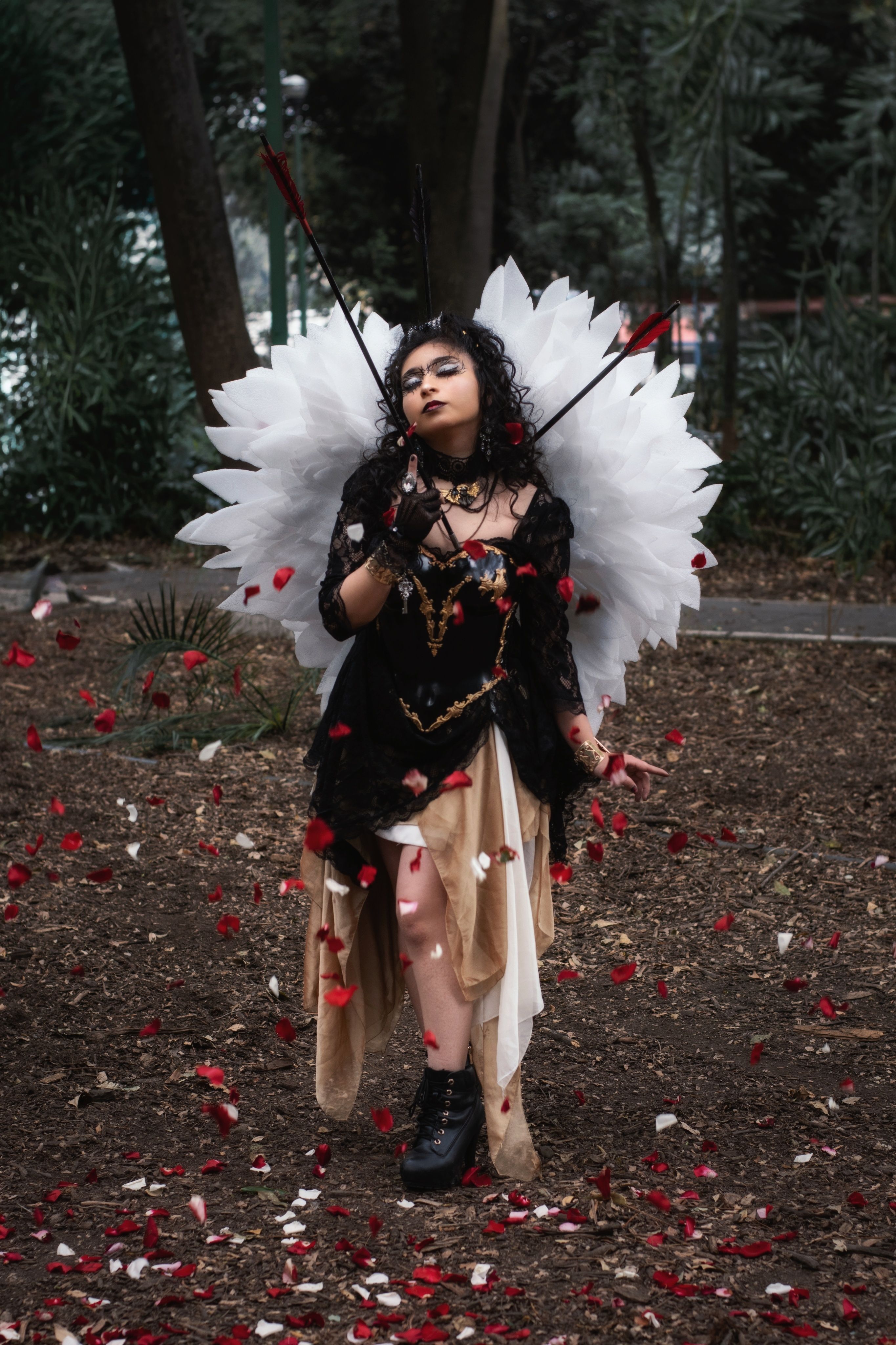 Person in a gothic angel costume with wings, outdoors surrounded by rose petals.