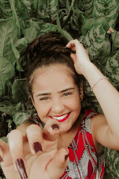 Happy woman lying in lush green foliage, reaching towards the camera with a joyful smile.