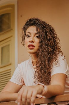 Young woman with curly hair in a relaxed pose inside an indoor setting, exuding confidence and style.