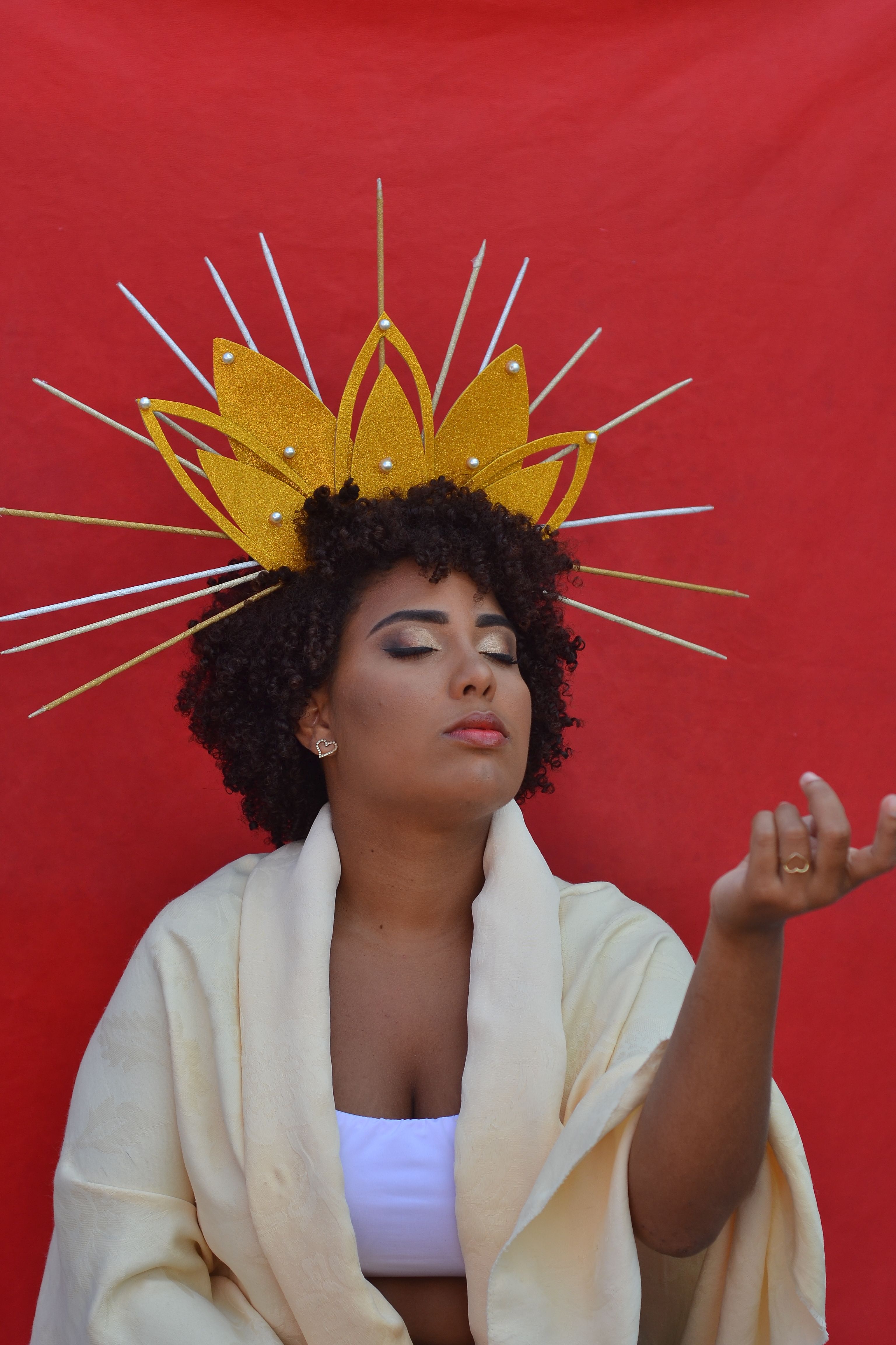 A woman wearing a radiant crown and white robe poses against a vivid red backdrop.