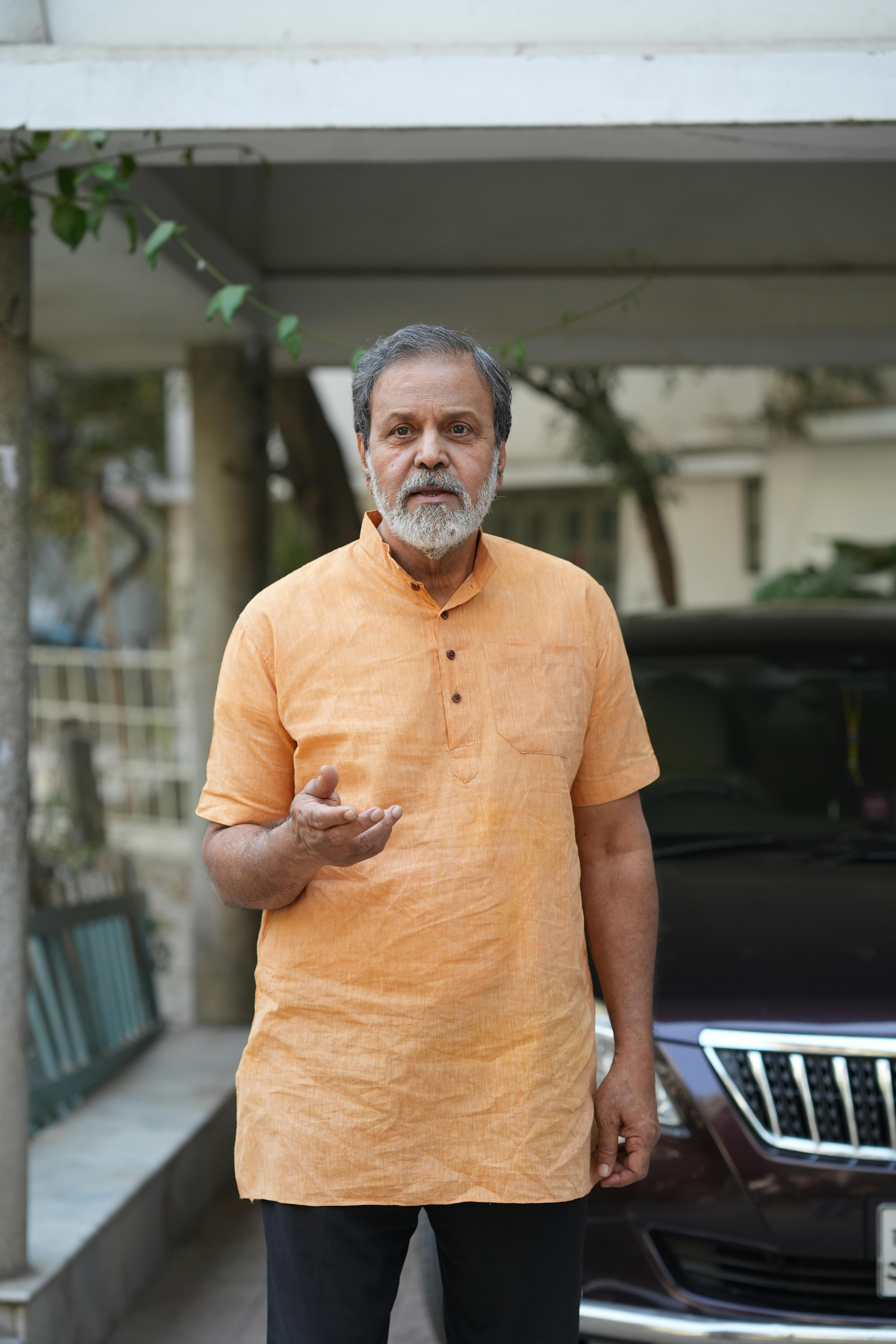 Elderly man in traditional orange kurta standing outside, next to a parked car.
