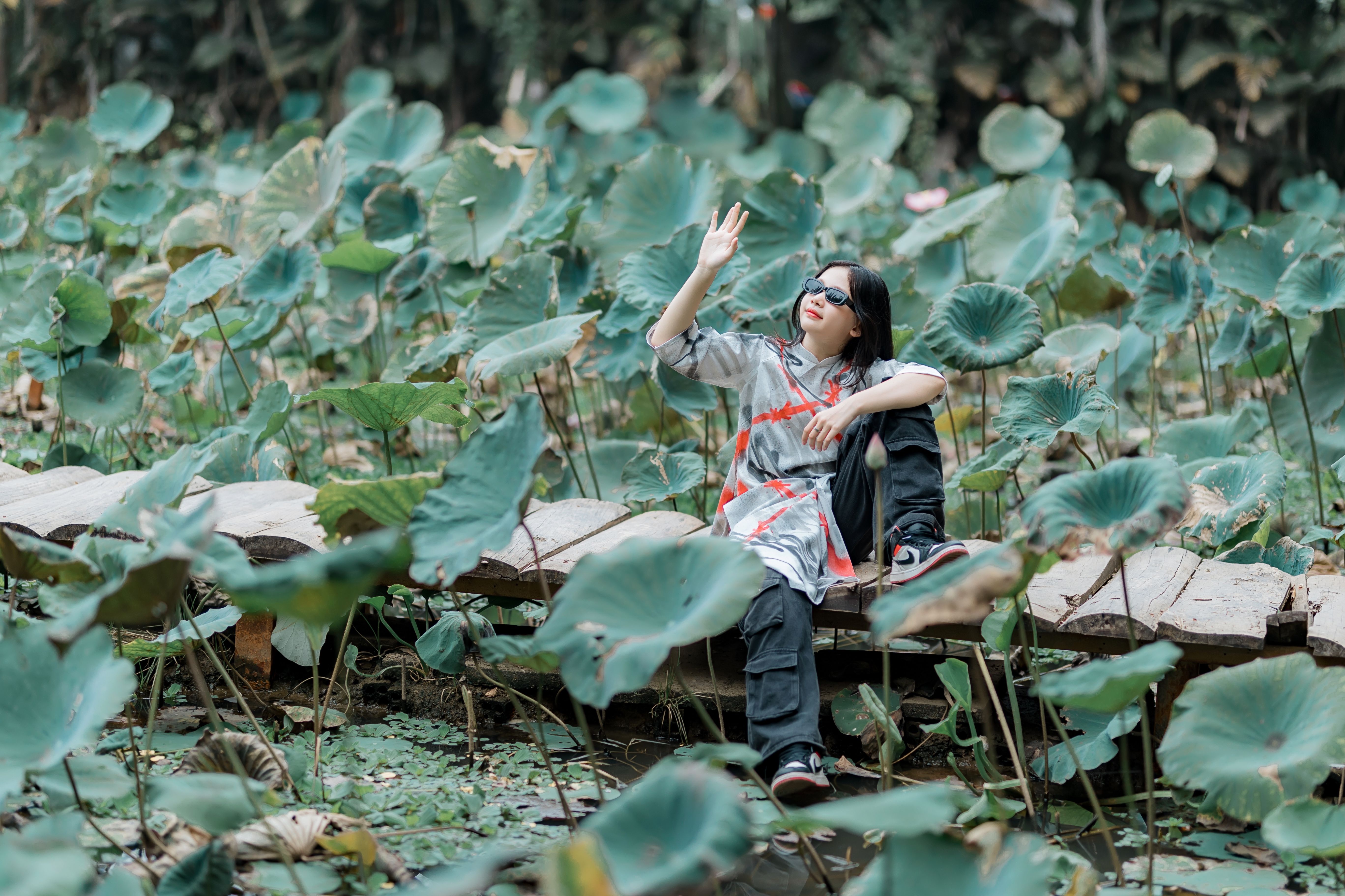 A woman in sunglasses relaxes on a wooden bridge surrounded by lush lotus leaves outdoors.