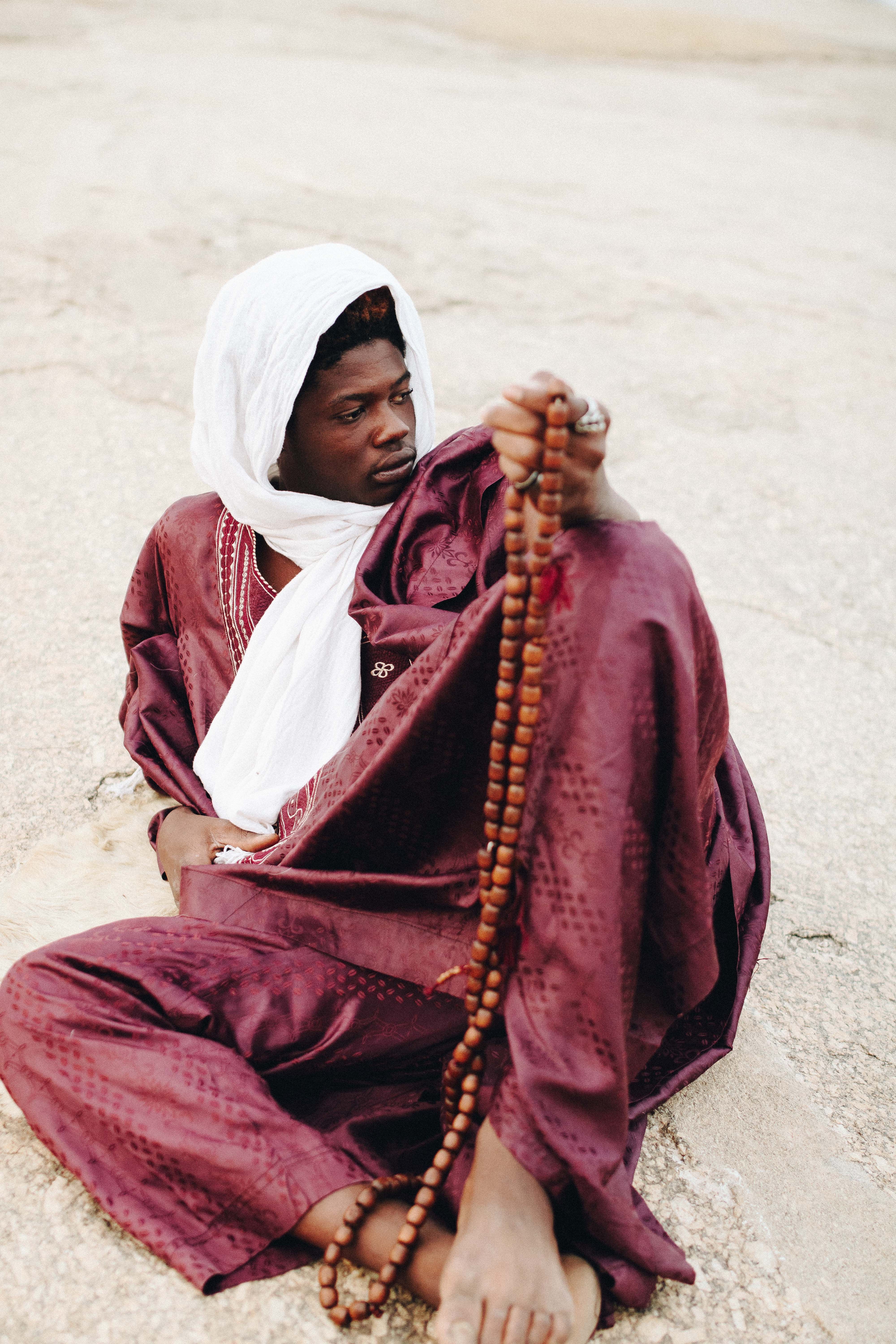 A young man in traditional attire holding prayer beads while seated on a sunlit surface outdoors.
