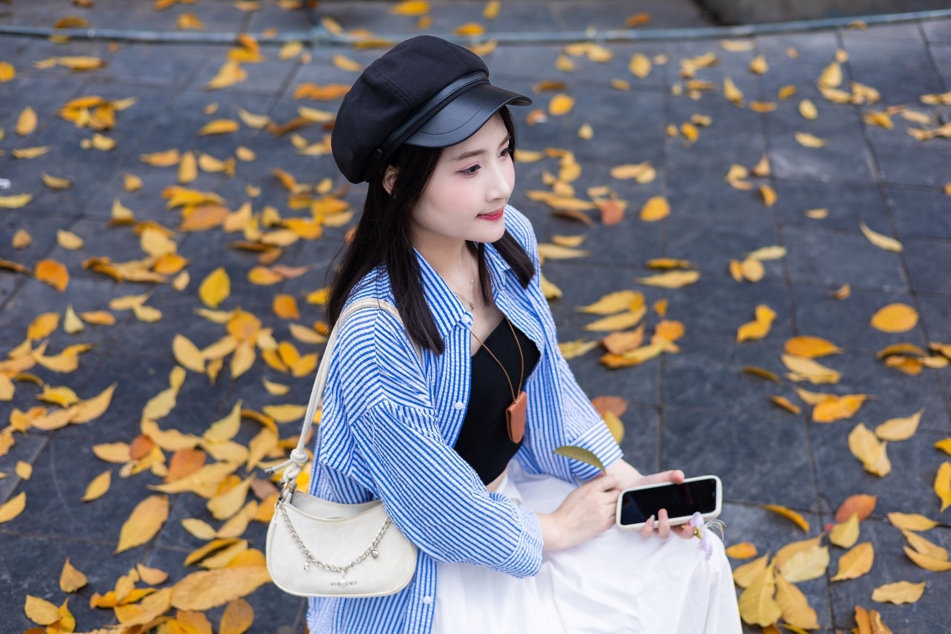 Young woman seated outdoors in Hanoi, holding a smartphone amidst fallen autumn leaves.