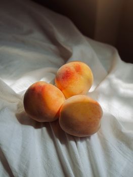 A trio of ripe peaches resting on a bed of soft white fabric, bathed in gentle sunlight.
