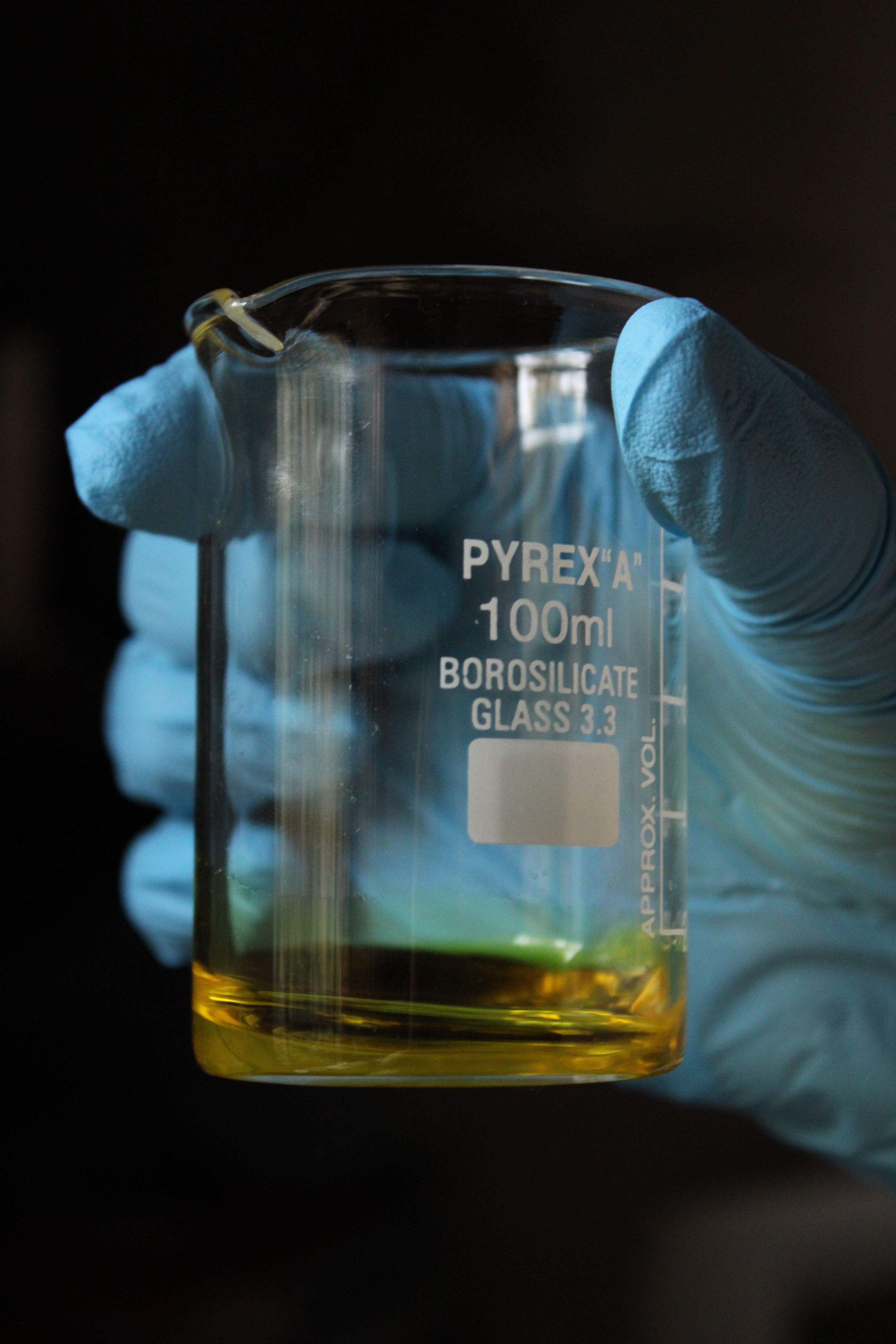 A beaker held by gloved hands in a lab setting, containing a yellow liquid.