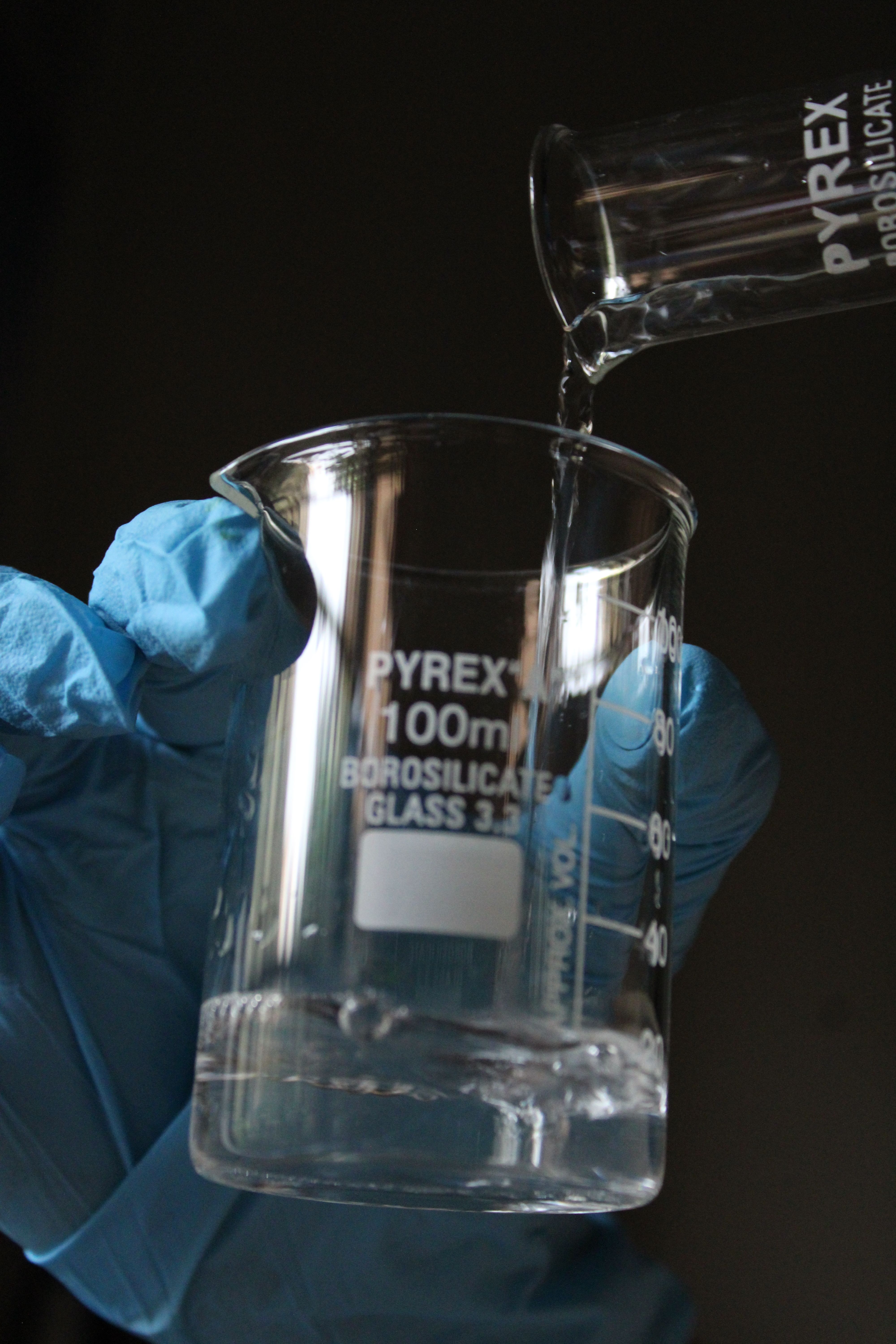 Close-up of a gloved hand pouring liquid into a Pyrex laboratory beaker.