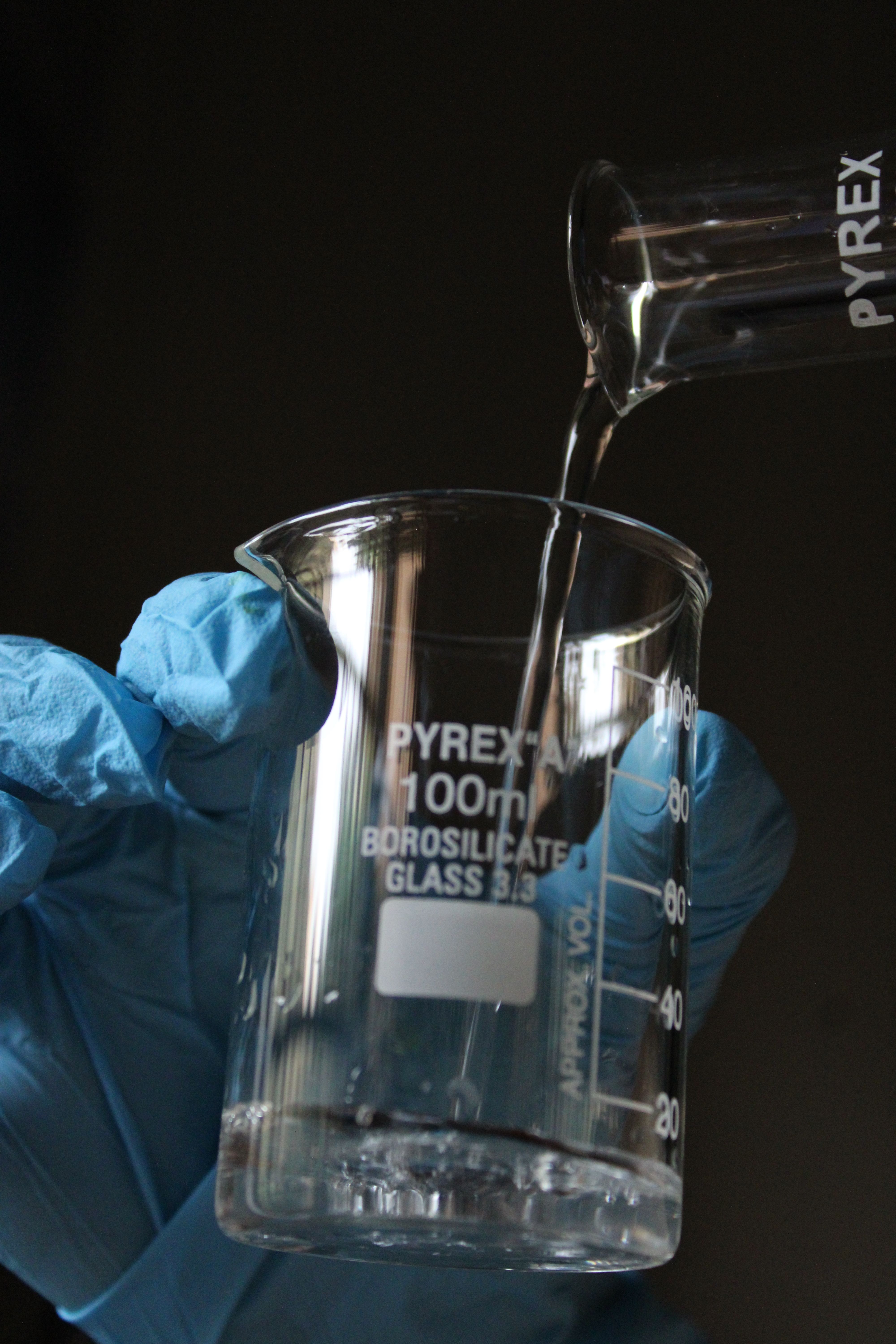 Close-up of a scientist's gloved hand pouring liquid into a Pyrex beaker.