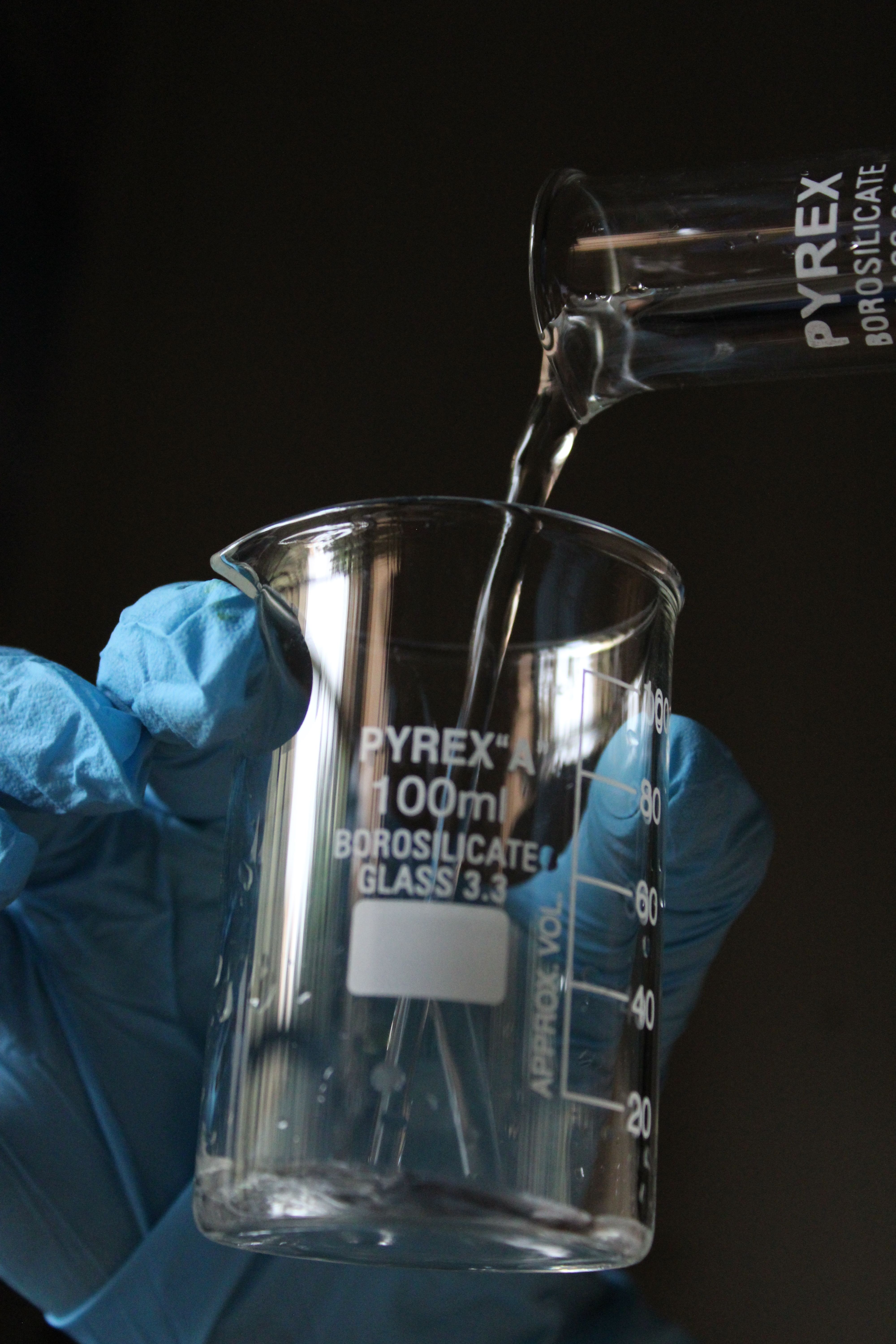 Gloved hand pouring liquid into a Pyrex beaker in a laboratory setting.
