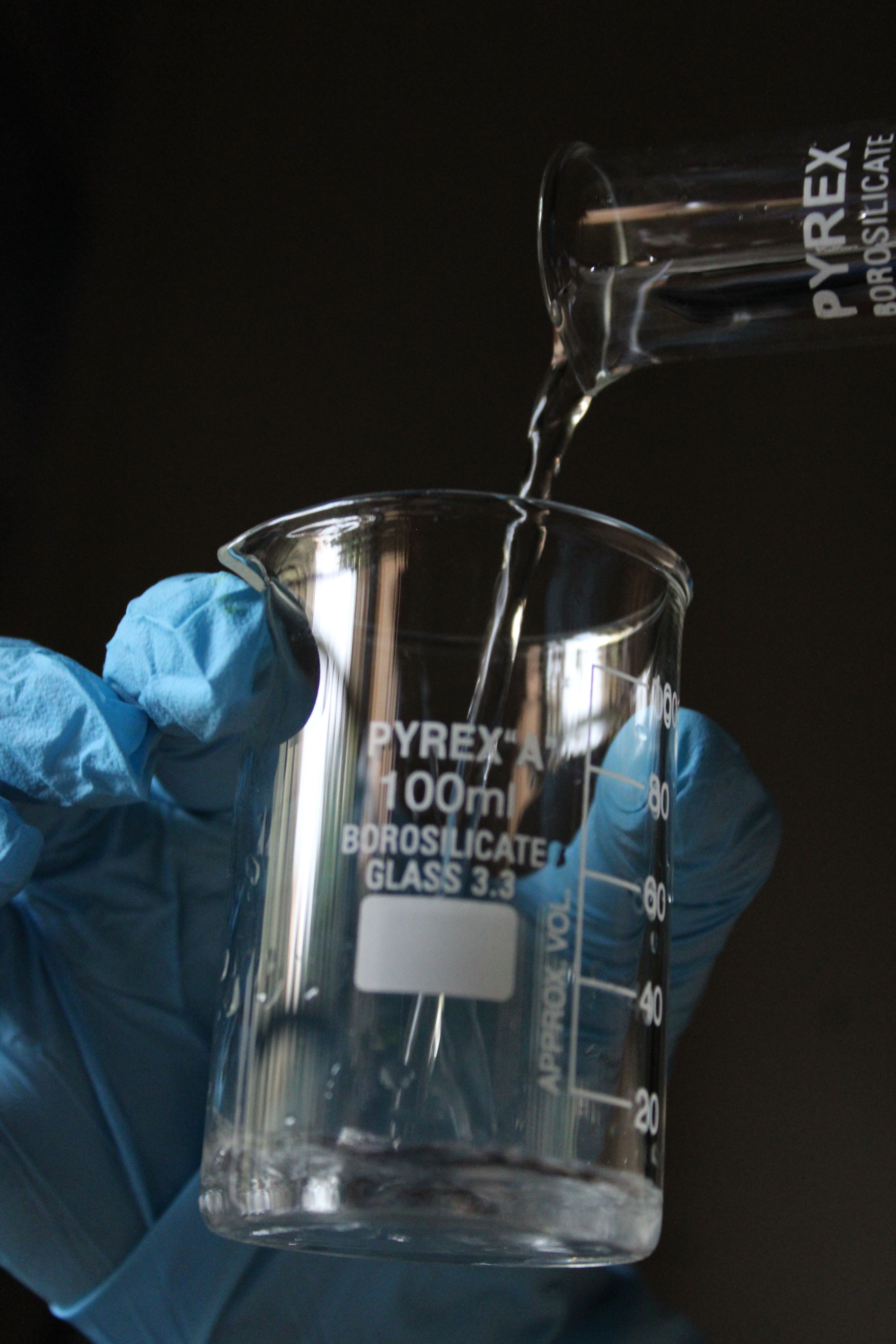Close-up of liquid being poured into a Pyrex beaker by a gloved hand.