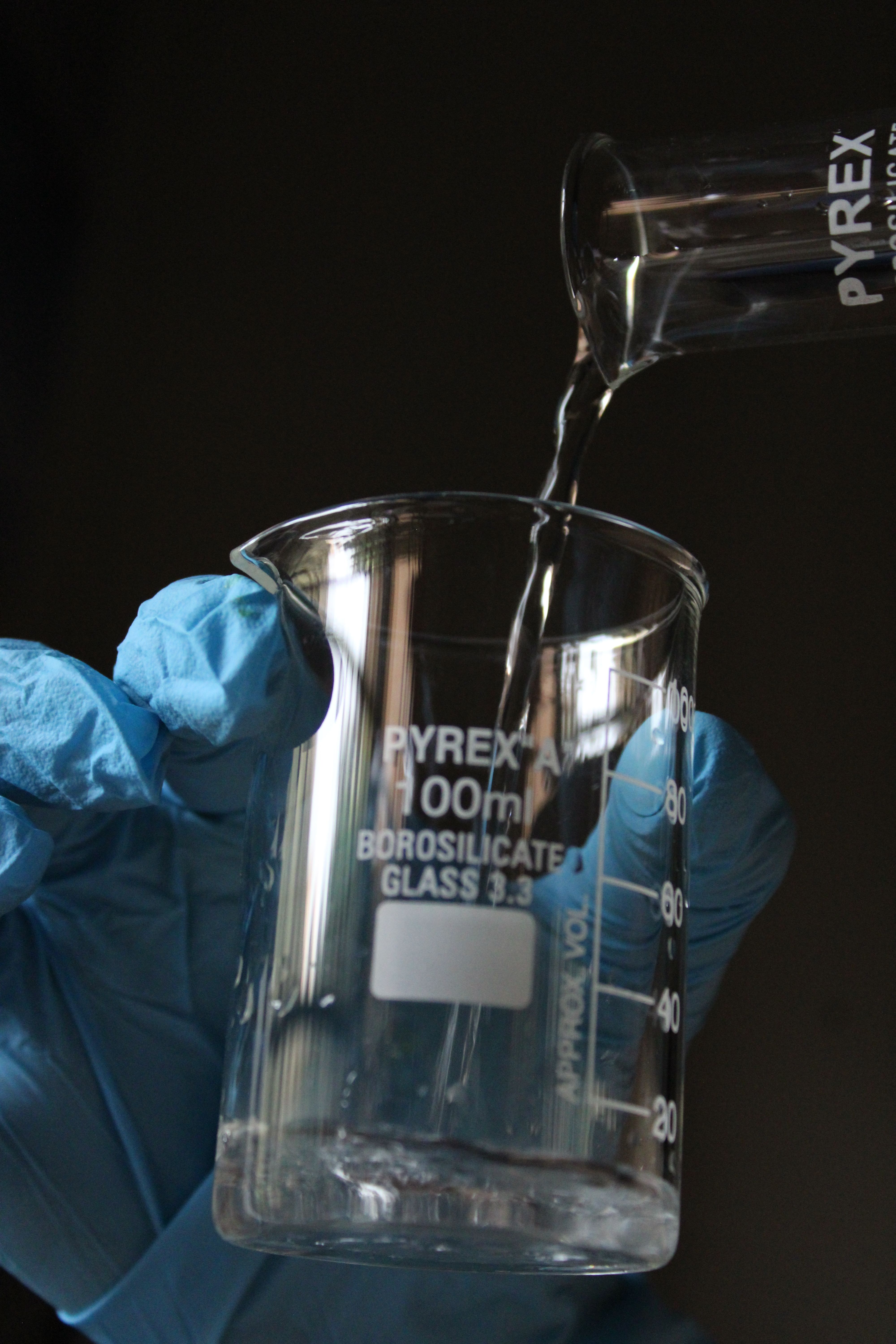 Close-up of a hand pouring liquid into a 100ml Pyrex beaker in a lab.