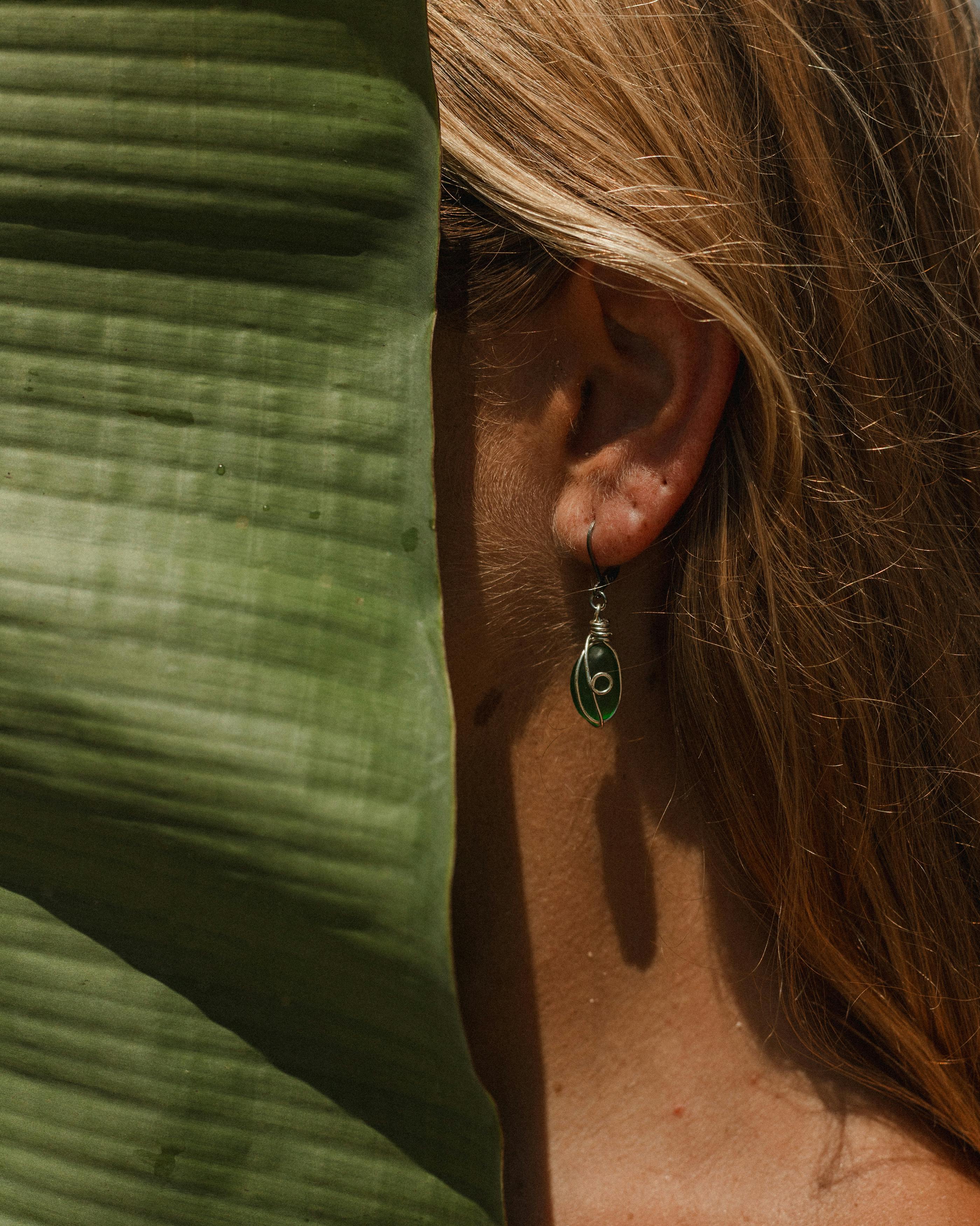 Free Close-up of a woman partially hidden by a large tropical leaf, highlighting a green earring. Stock Photo