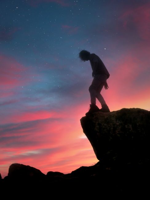 Silhouette of a person standing on a rock under a starry sky at sunset with pink hues.