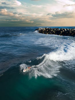 Drone shot capturing a surfer riding waves near a rocky breakwater under a cloudy sky.