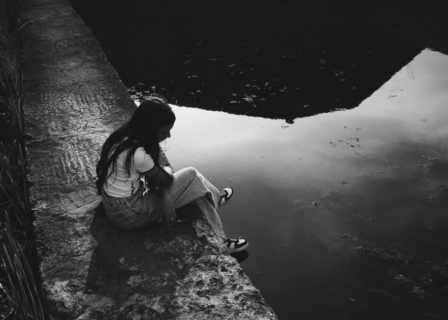Black and white photo of a woman sitting pensively by the water, capturing a reflective moment.