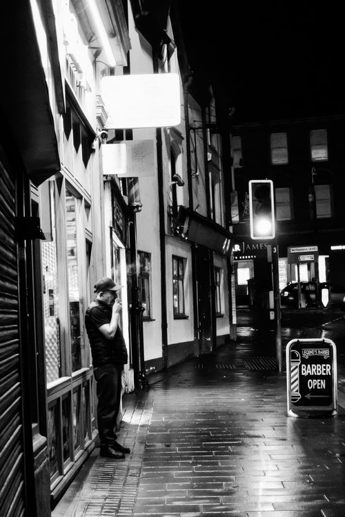 Free A man smoking outside a shop on a rainy night in a dimly lit urban street. Stock Photo