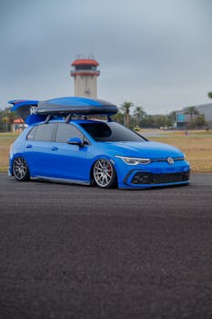 A sleek blue hatchback car with a roof box parked near an airport with control tower in view.