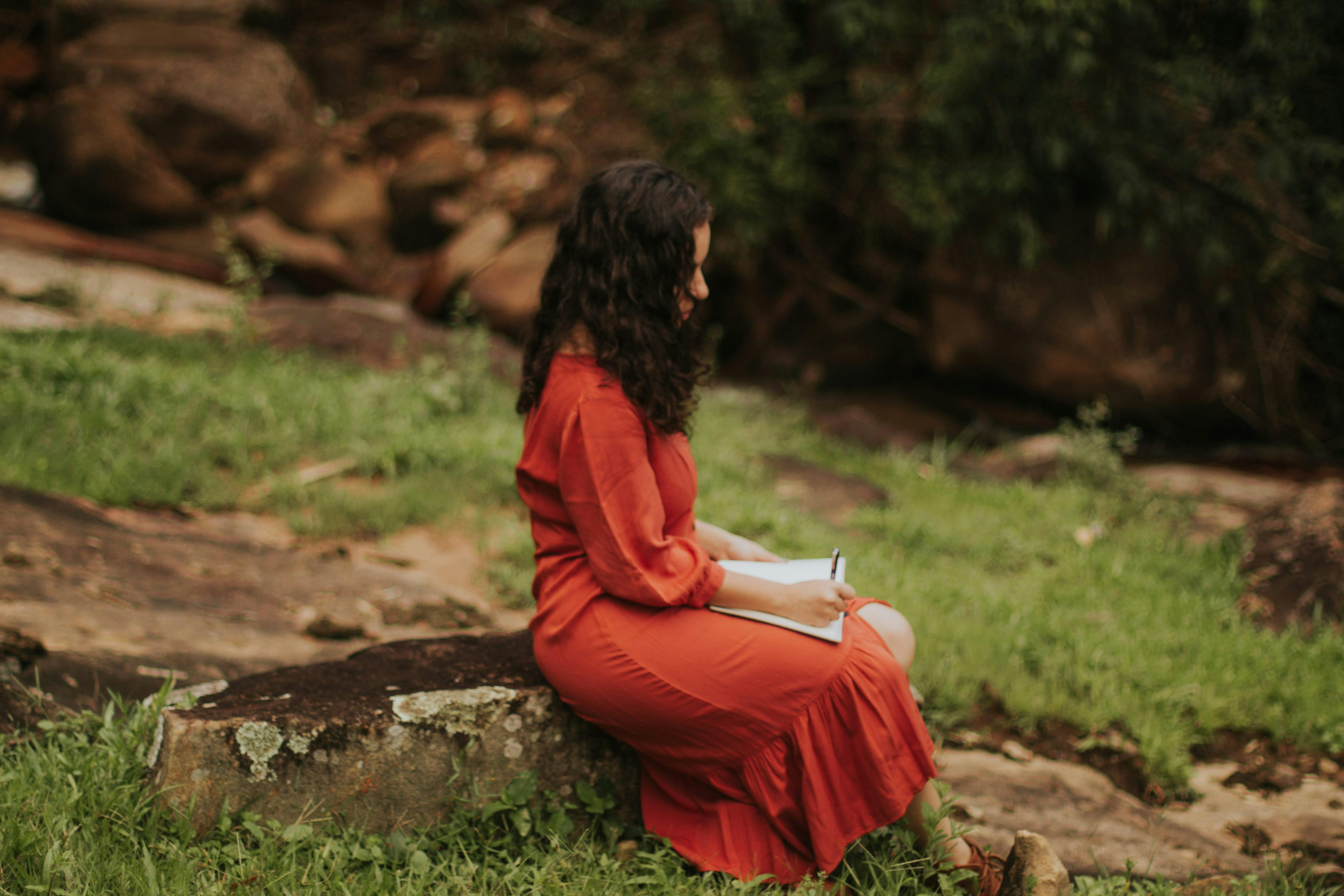 A woman sits on a rock, writing in a notebook in a serene outdoor setting.