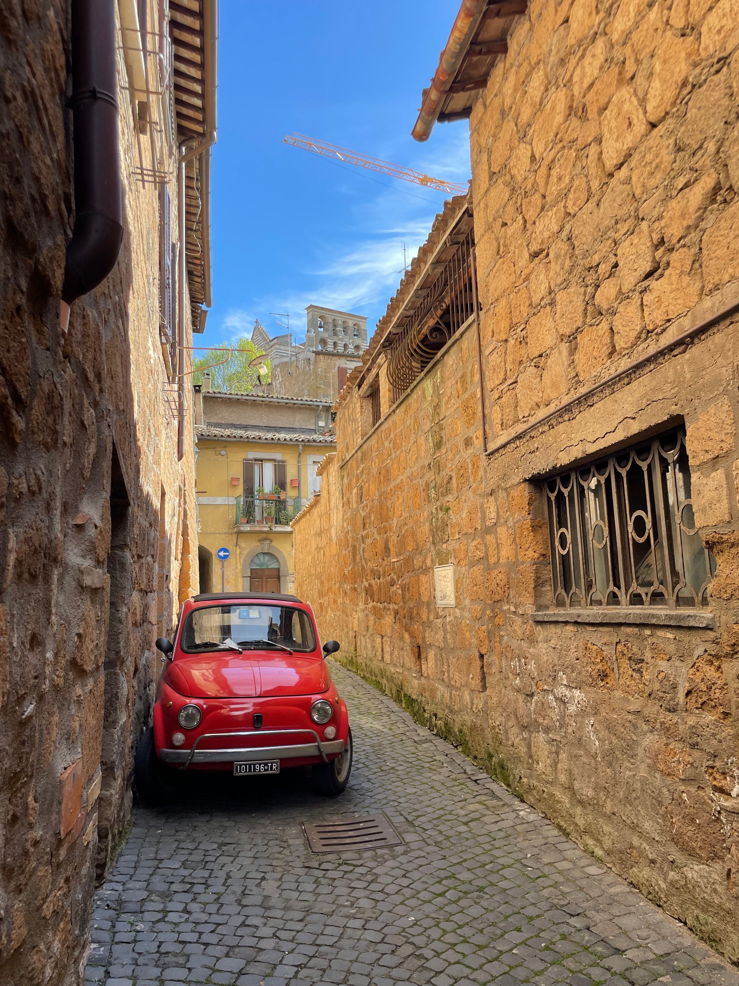 Explore a quaint alley in Orvieto, Italy, featuring a classic red Fiat 500 parked against rustic stone walls.