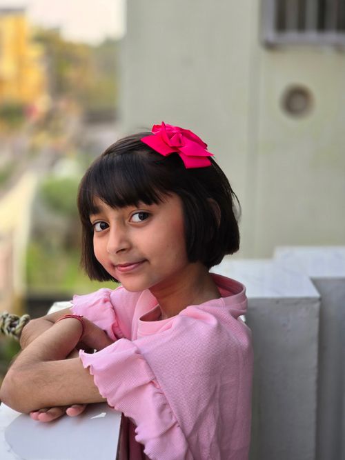 Free Smiling girl with a pink bow posing on a balcony, showcasing innocence and joy. Stock Photo