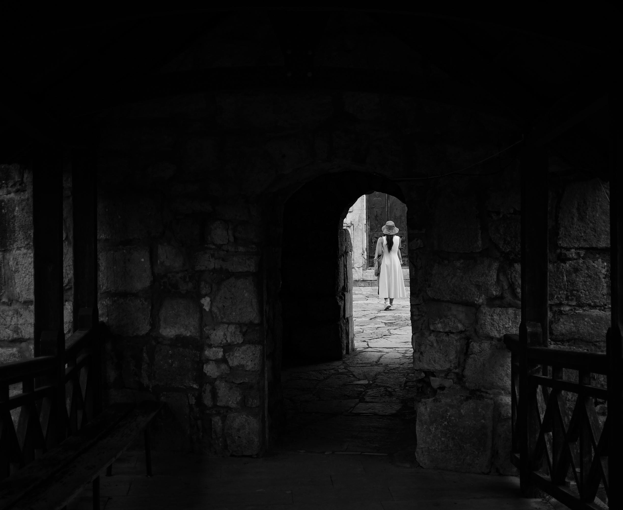 A woman in a dress and hat walks through an ancient stone archway into the courtyard.